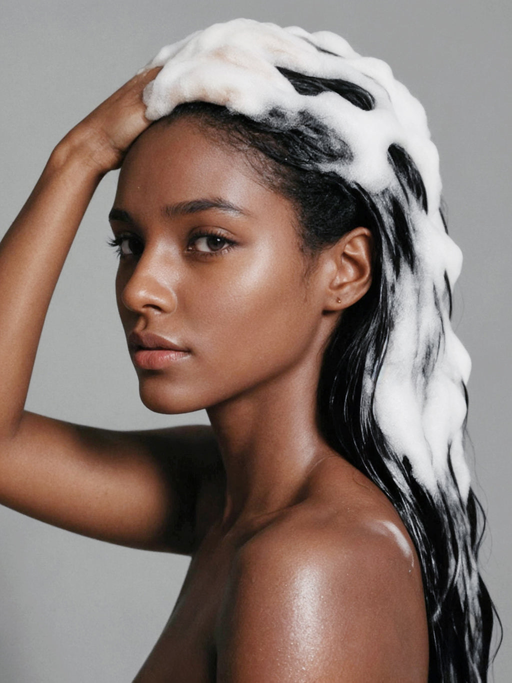 A close-up portrait of a woman with dark hair covered in thick white shampoo lather, posing against a neutral gray background.